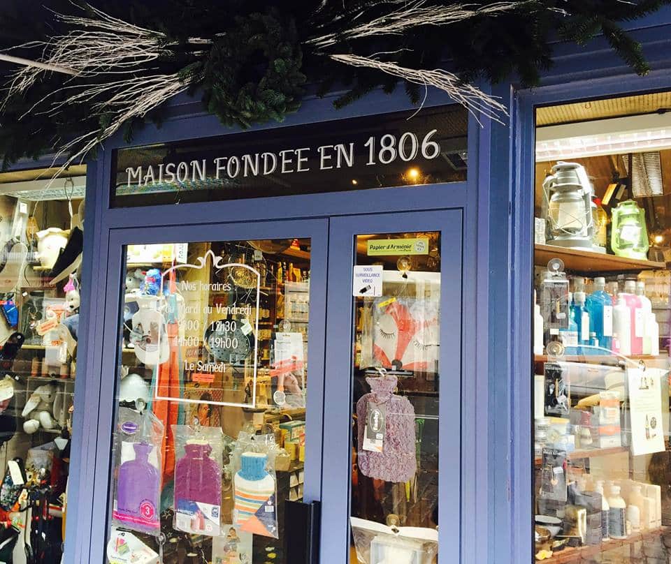 Maison fondée en 1806 : Boutique Parisienne Ancienne Façade d'une boutique française "Maison Fondée en 1806" avec horaires inscrits : Lun-Ven 10h00-12h30/14h00-19h00, Le Samedi. Vitrines remplies de produits et décorations hivernales.